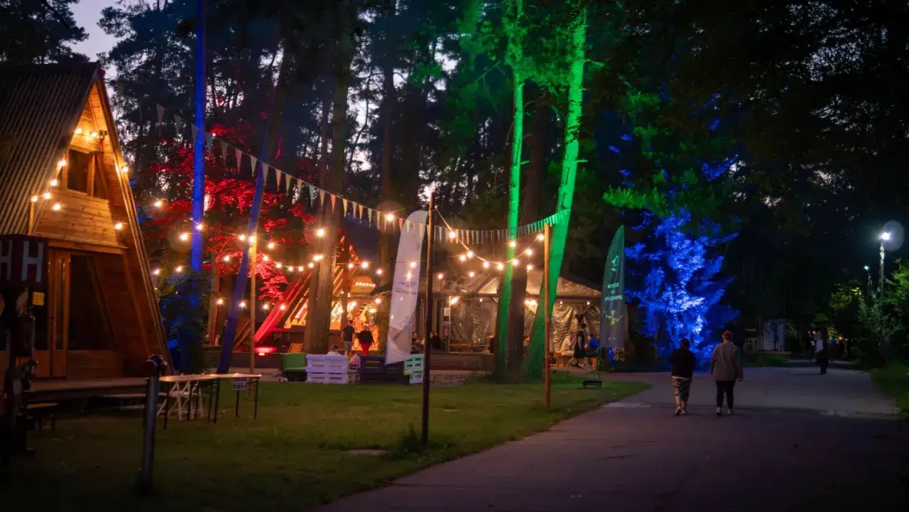 Nocny festiwal w lesie, oświetlone domki i drzewa kolorowymi światłami. To idealne miejsce na nocleg po dniu pełnym wrażeń na Pyrkonie.