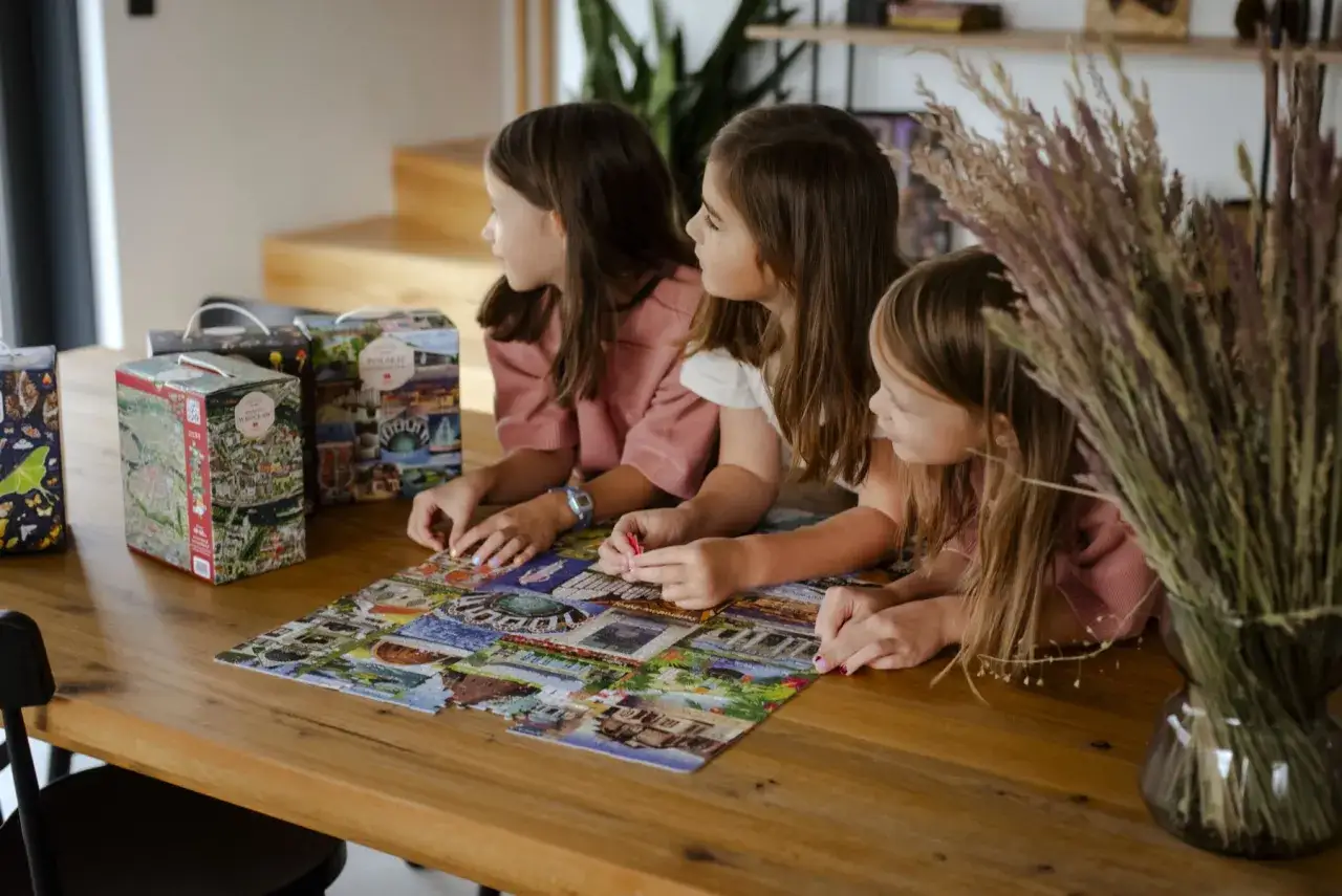 Trzy dziewczynki układają puzzle, ćwicząc spostrzegawczość i cierpliwość. Obok pudełka z widokiem na Wrocław.