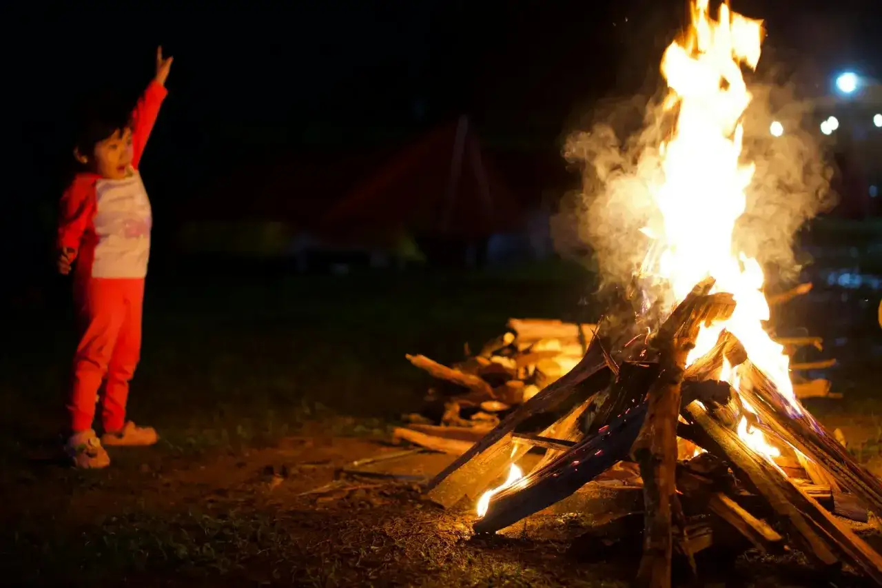 Dziecko w pomarańczowych spodniach i bluzie z podniesioną ręką obserwuje płomienie ogniska. Wesołe zabawy przy ognisku.