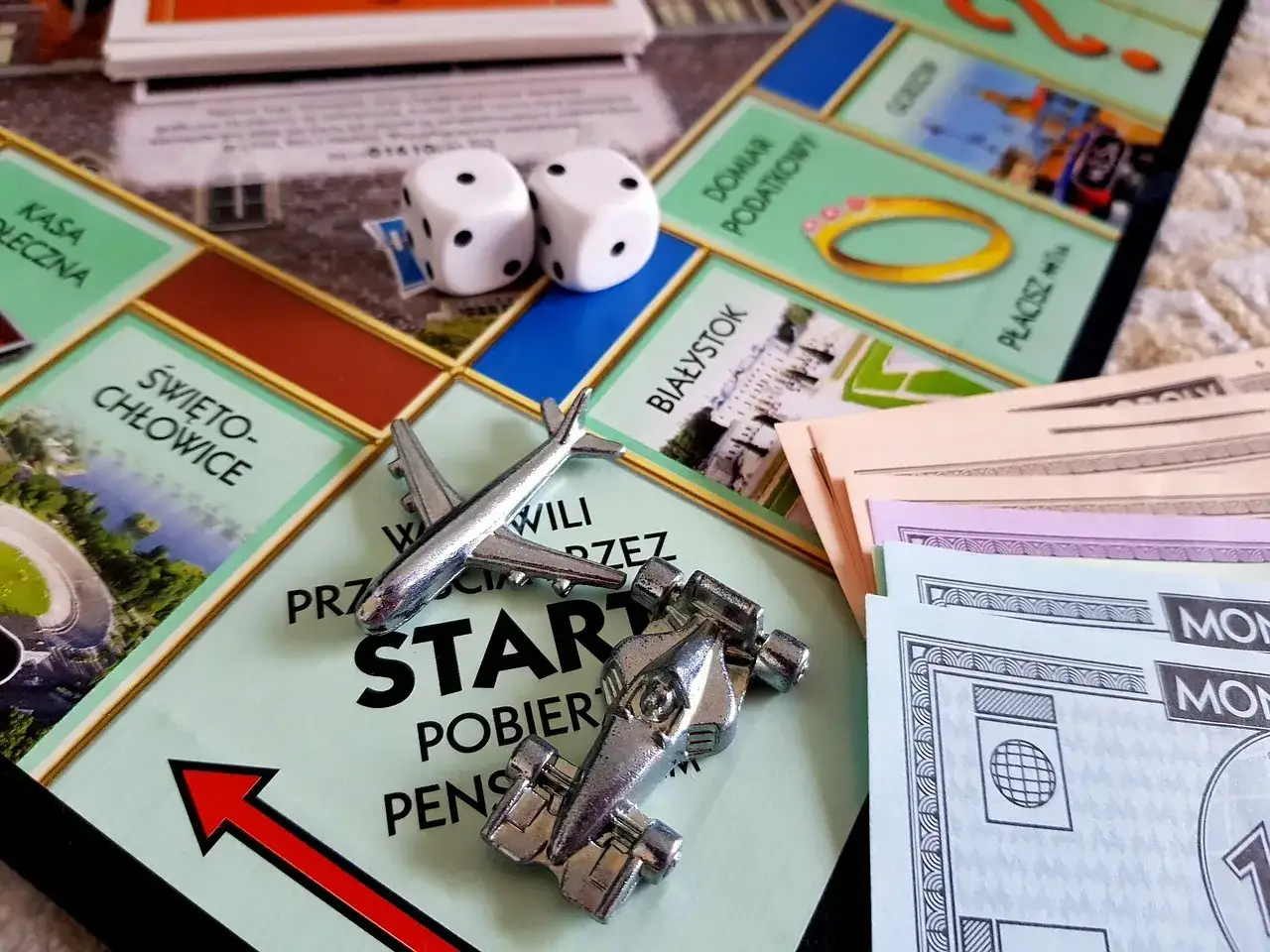 Kostki, samolot i bolid na planszy Monopoly. Różne rodzaje pól, pieniądze i pionki sugerują rozgrywkę w tę klasyczną grę.