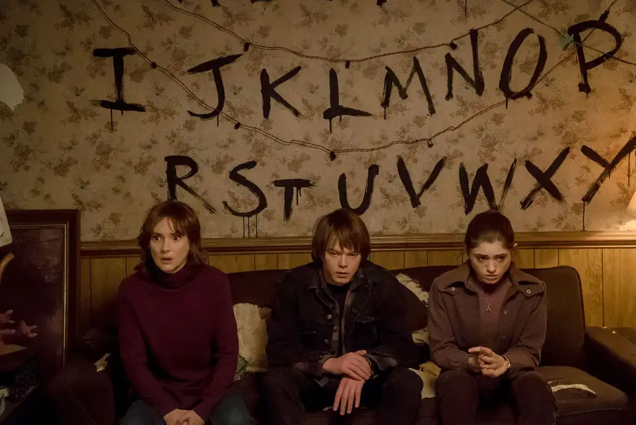 Joyce Byers i jej synowie, Jonathan i Nancy, siedzą przed ścianą z namalowanymi literami i lampkami, kluczowy element w Stranger Things.