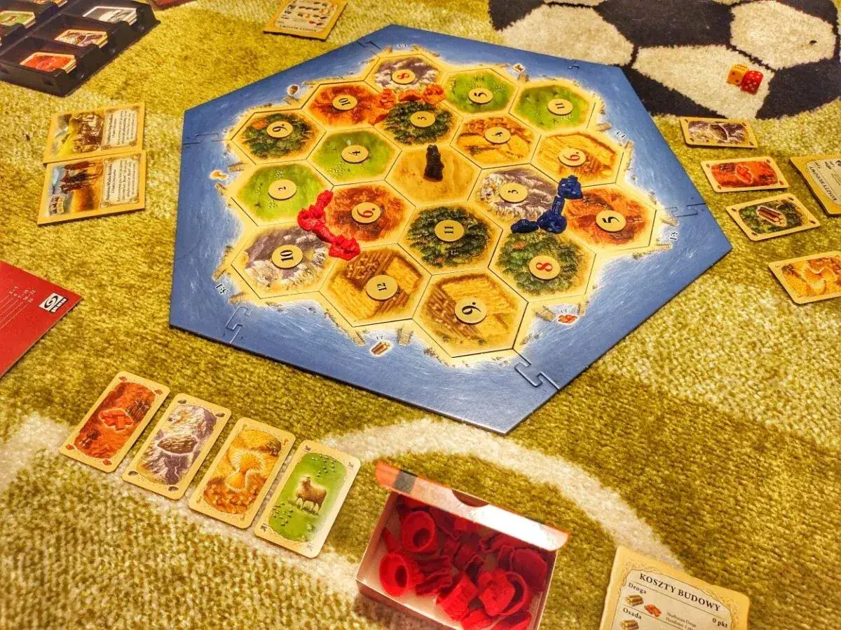 Plansza do gry Catan, karty surowców i pionki. Rozgrywka w Catan instrukcja w tle.