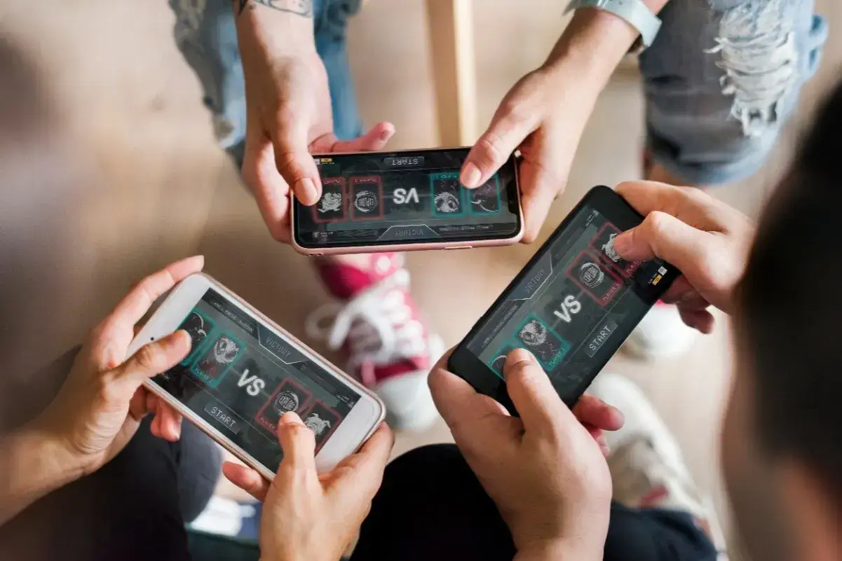 Trzy osoby grają w gry grupowe na telefonie. Na ekranach widać planszę z napisem