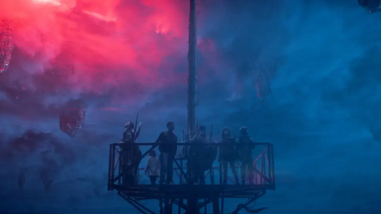 Grupa bohaterów na platformie obserwuje surrealistyczny krajobraz z unoszącymi się skałami. To scena jak ze stranger things scenariusz.