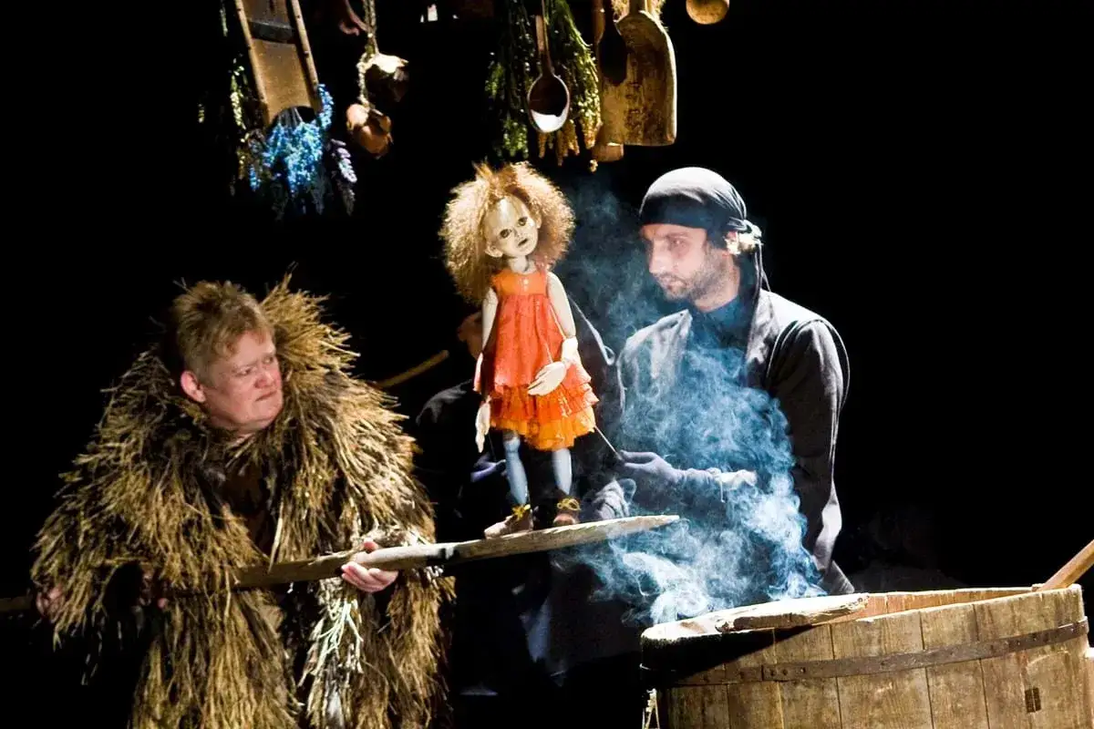 Baba Jaga patrzy, jak kukła w pomarańczowej sukience tańczy w dymie. Obok niej mężczyzna w słomianym płaszczu.