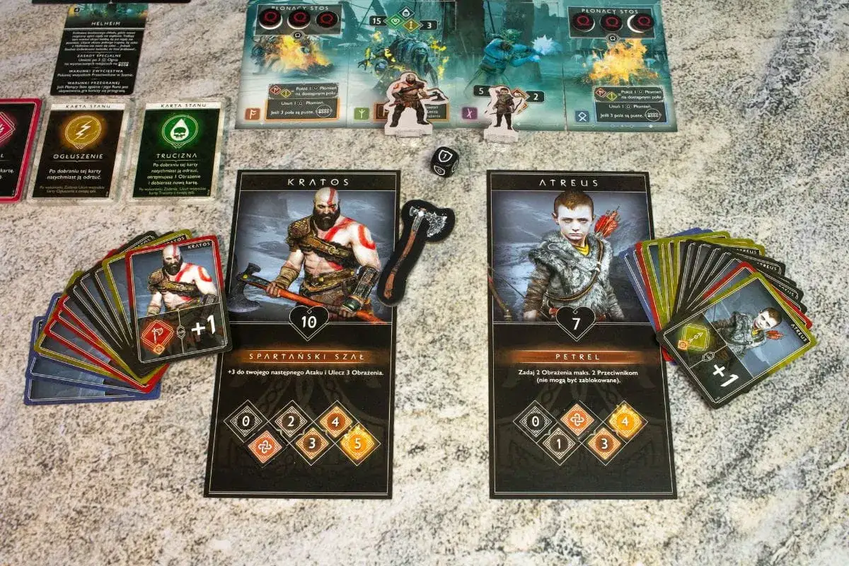 Karty Kratos i Atreus z gry planszowej. Obok stosy kart, kostka i żetony. To ja go tnę dodatki, przygotowując się do walki.