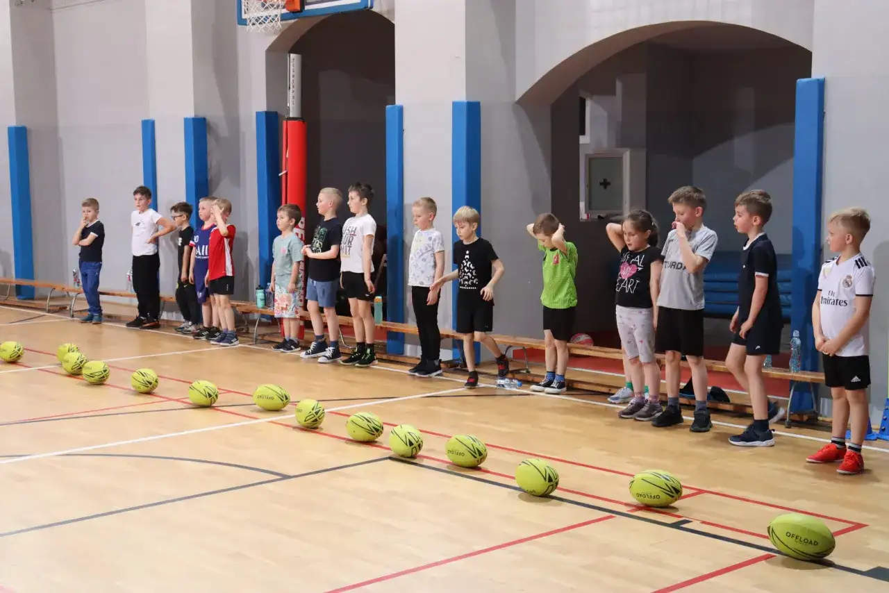 Dzieci w hali sportowej czekają na śmieszne konkurencje sportowe. Przed nimi leżą żółte piłki.