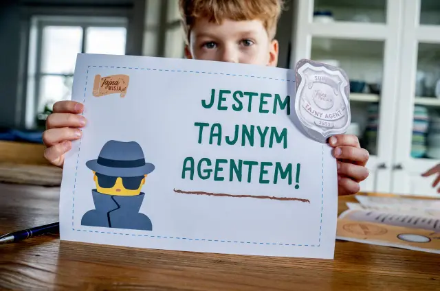 Chłopiec trzyma kartkę z napisem "Jestem tajnym agentem!" i odznaką. Idealne do gier escape room.